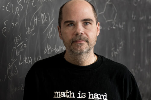 Professor Alan Koch stands in front of a chalkboard with math equations in the background.