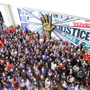 a large group of students wearing purple shirts stand in front of a wall mural encouraging justice