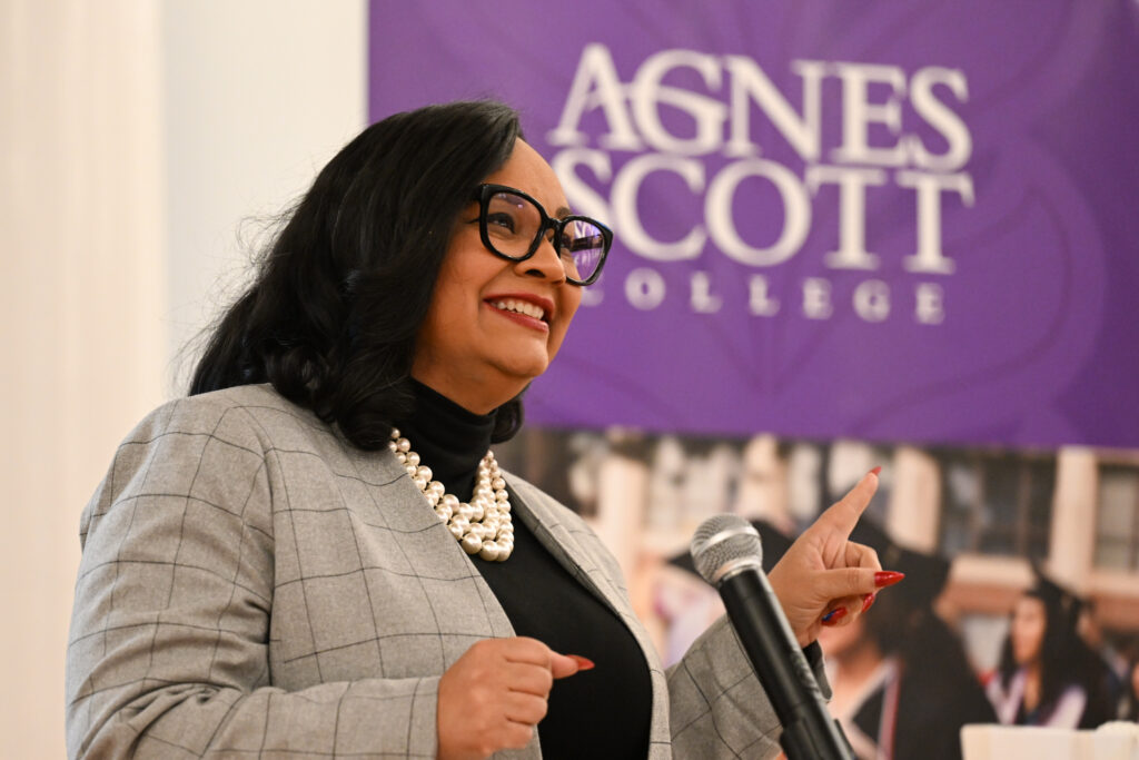 Congresswoman Nikema Williams speaks into a microphone. The congresswoman points with her left hand in the direction of an Agnes Scott College banner.