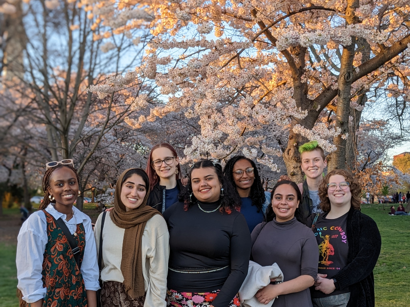 Eight students pose for a photo in front of dozens of cherry trees. The pinkish white flowers are in full bloom. 
