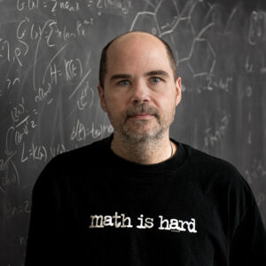 Professor Alan Koch stands in front of a chalkboard with math equations in the background.