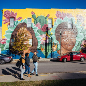 Three students walk on a sidewalk. A brightly colored mural is overlaid on a brick building behind them.