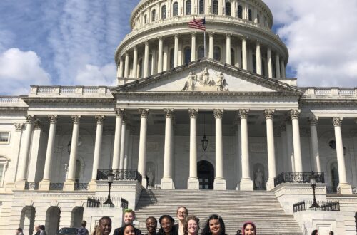 A group of students stand in front of the United States Capitol Building. A bright blue sky full of fluffy clouds sits over head.