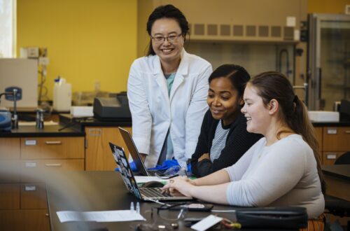 A faculty member in a white medical coat looks on as two students discuss something in front of a laptop