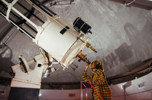 a student stands on a ladder while looking from right to left into a large white telescope