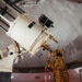a student stands on a ladder while looking from right to left into a large white telescope
