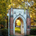Fall yellow leaves are illuminated by the sun behind a welcome arch at the front of a college campus.