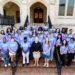 A group of graduate students wearing powder blue shirts pose for a photo with a college president in front of a gothic brick building