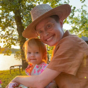 A women with a brimmed hat smiles in the light of a sunset while holding a young child.