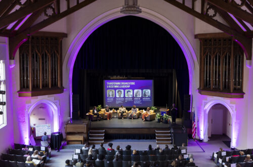A photo from a balcony of five people sitting in chairs on a stage. Purple stage lighting surrounds them.