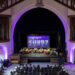 A photo from a balcony of five people sitting in chairs on a stage. Purple stage lighting surrounds them.