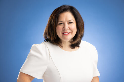 A woman poses for a headshot in front of a blue background while wearing a white dress.