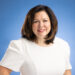 A woman poses for a headshot in front of a blue background while wearing a white dress.