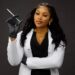 A confident woman with long, wavy black hair poses against a dark background. She is wearing a white lab coat over a black outfit, gold jewelry, and black gloves. She holds a syringe in her right hand, raised slightly, and gazes directly at the camera with a poised expression. Her left arm is crossed over her body.